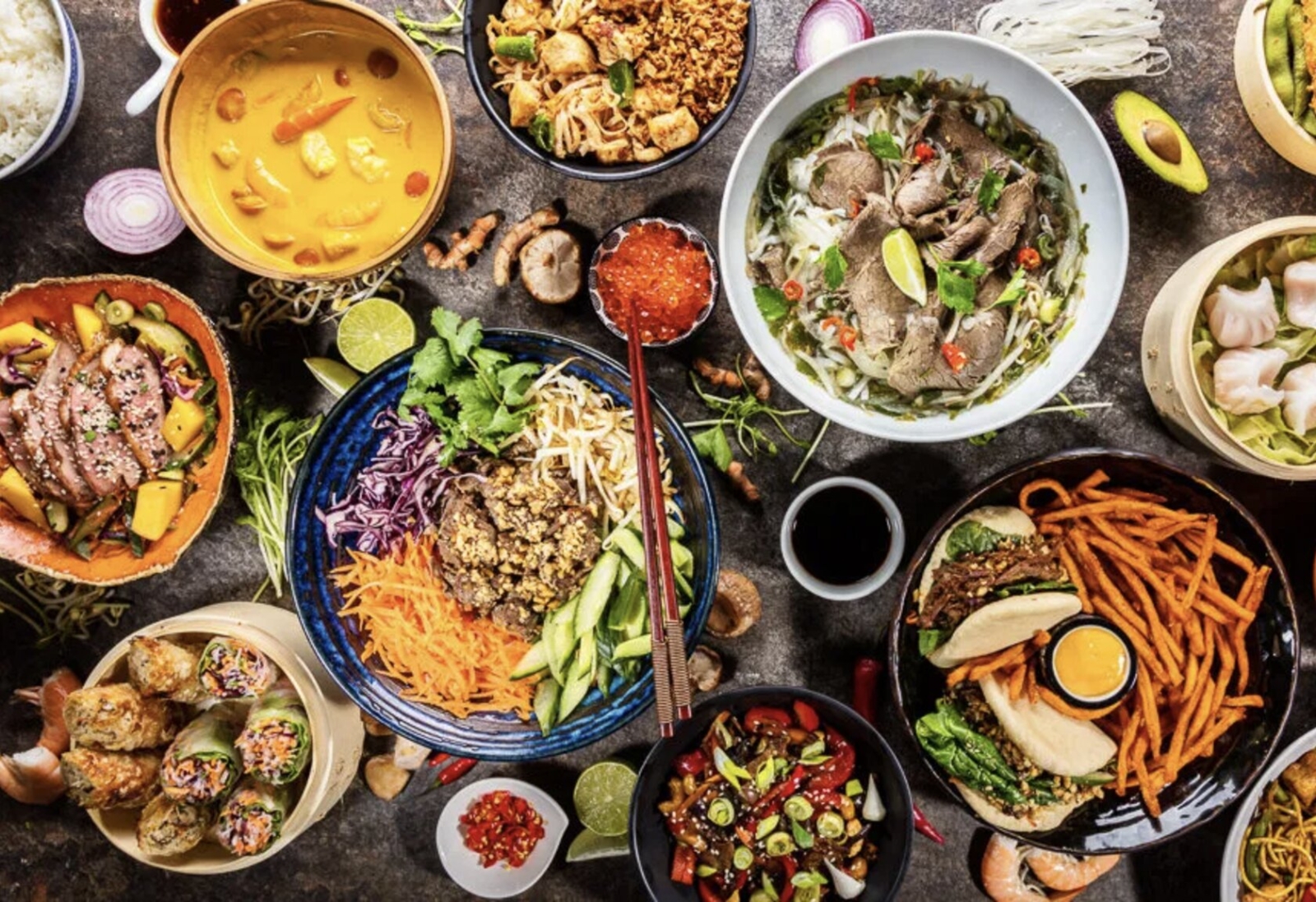 A diverse table spread showcasing vibrant Asian fusion dishes, including sushi rolls, noodle bowls, curries, and fresh vegetables.